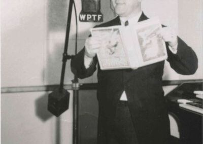 Historic black-and-white photo of a man in a top hat standing at a WPTF microphone, holding an illustrated booklet while performing in the studio.