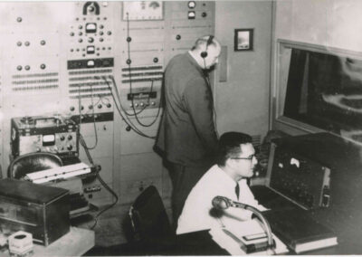 Black-and-white photo of two WPTF engineers in a room lined with tall audio equipment racks, monitoring levels and managing broadcast gear.