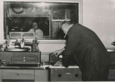 Historic studio image of an engineer leaning over disc-cutting equipment while another engineer observes through a control-room window.