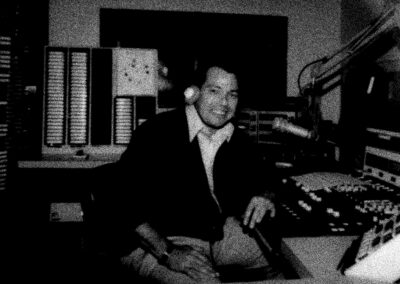 Black-and-white photo of a smiling WPTF radio announcer wearing headphones and sitting at a broadcast console surrounded by audio equipment and tape racks.