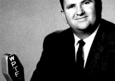 Formal black-and-white portrait of a man in a suit seated beside a vintage WPTF broadcast microphone.