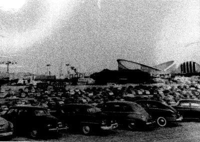 Black-and-white photo showing a crowded parking lot filled with mid-century automobiles and large fairground-style structures in the background.