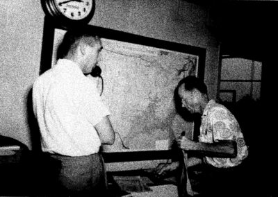 Black-and-white photo of two men in a WPTF office examining a large wall map; one speaks into a phone while the other takes notes.